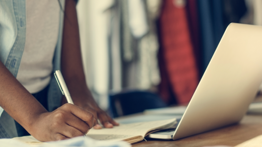 A person stands at a table, writing in a notebook with one hand while the other rests nearby. An open laptop, folded clothes, and blurred clothing racks suggest an environment focused on Retail Operations Consulting.