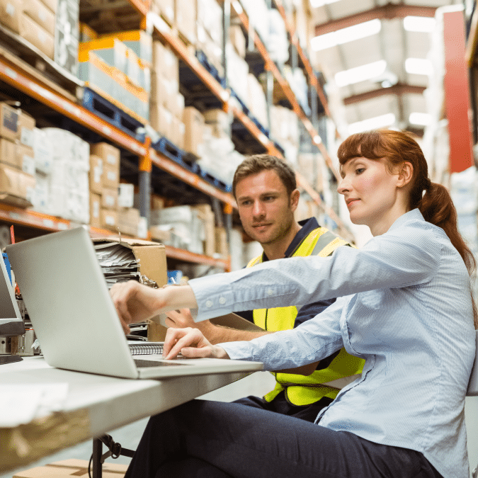 Two workers, one in a safety vest and the other in business attire, sit at a desk using a laptop in a warehouse filled with shelves of boxes and supplies.