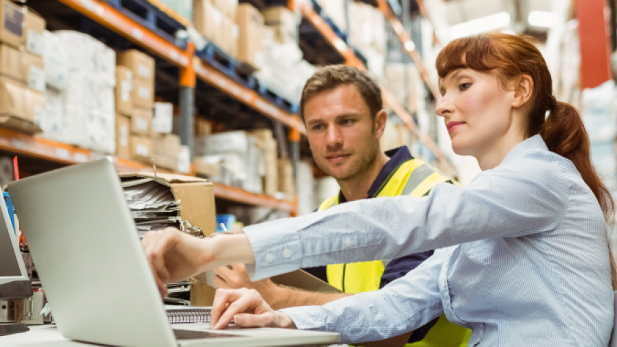 Two workers in a warehouse sit at a table with a laptop, discussing something on the screen. One wears a safety vest and the other is dressed in business attire. Shelves stocked with boxes are visible in the background.