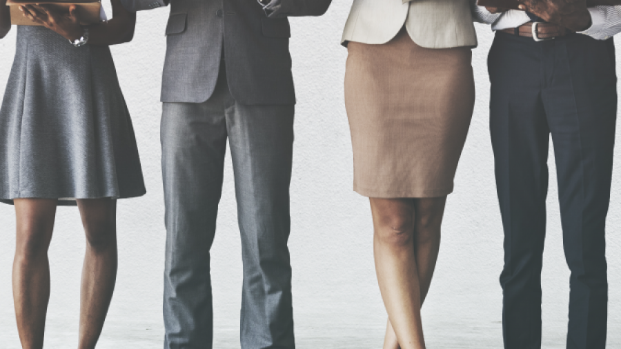 Four professionally dressed people stand side by side against a plain background, holding and reading documents or tablets, with their faces out of frame.