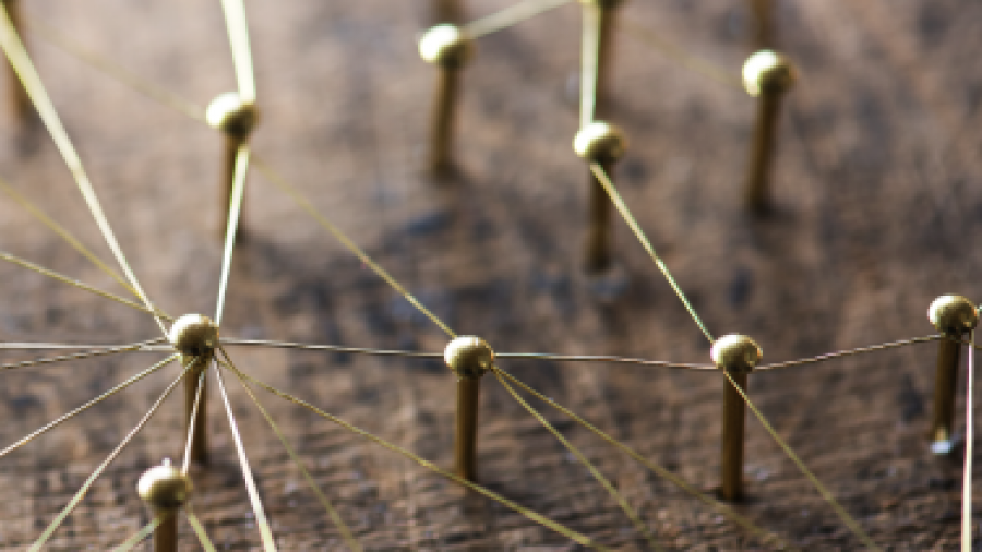 Close-up of brass pins connected by thin gold threads on a wooden surface, forming a network or web pattern, symbolizing connections or communication.