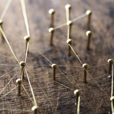 Close-up of brass pins connected by thin gold threads on a wooden surface, forming a network or web pattern, symbolizing connections or communication.