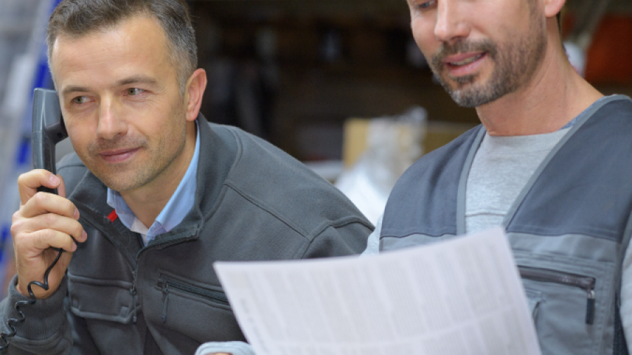 Two men in work uniforms sit in a warehouse. One is talking on the phone while the other reviews a document. Shelves with boxes and packages are visible in the background.