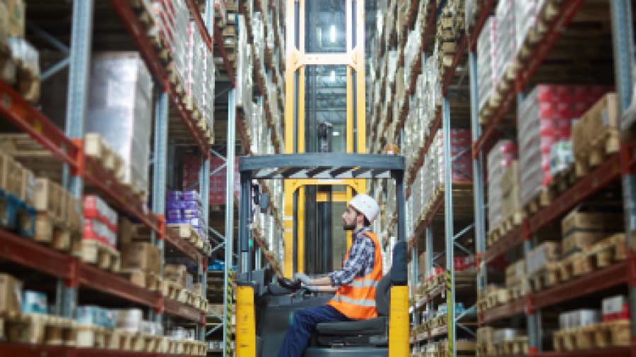 A worker wearing a hard hat and safety vest operates a forklift in a warehouse aisle lined with tall shelves stocked with various boxes and goods.