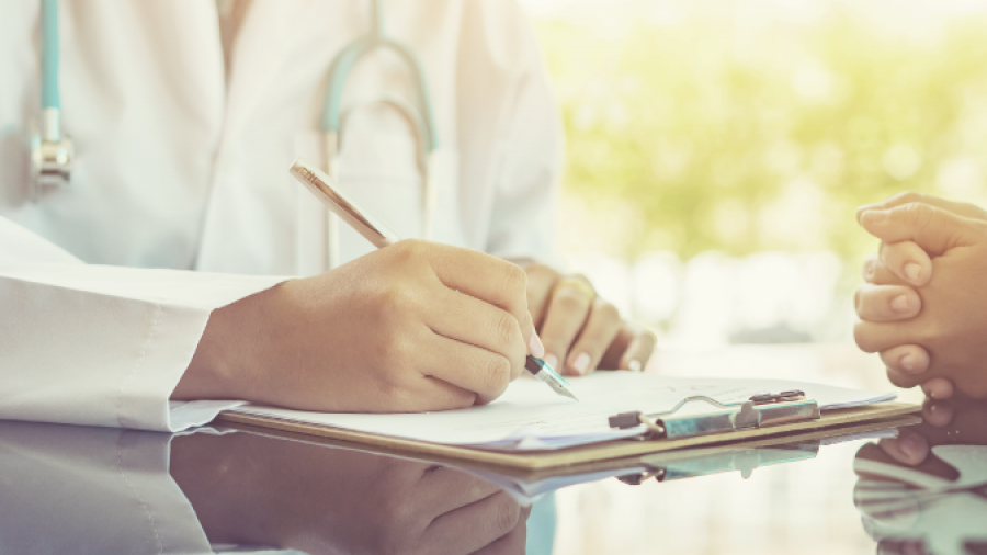 A doctor in a white coat holding a clipboard and writing notes, while another person sits across the desk holding the doctors hand, suggesting a comforting or supportive interaction.