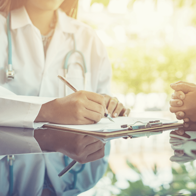 A doctor in a white coat holding a clipboard and writing notes, while another person sits across the desk holding the doctors hand, suggesting a comforting or supportive interaction.