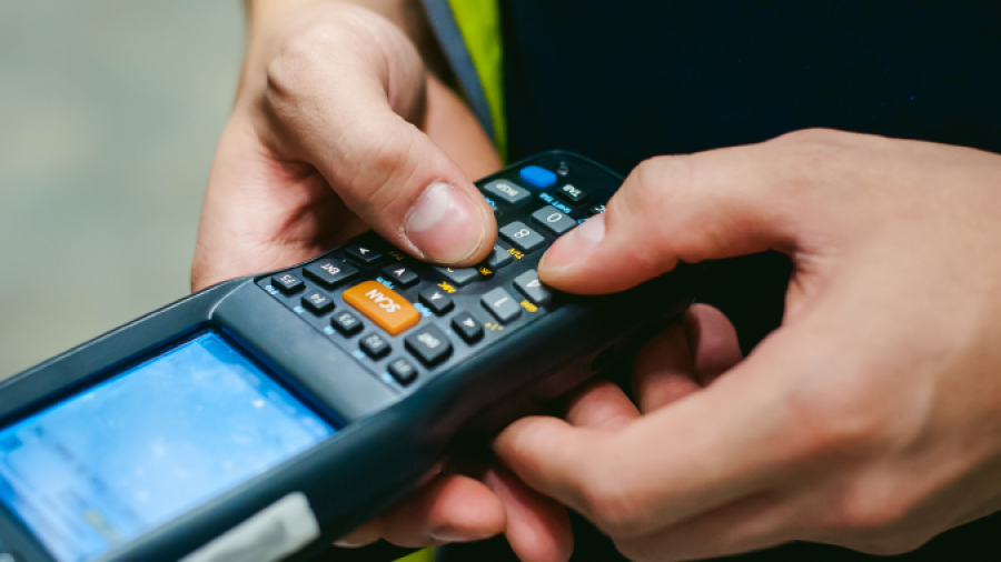 A person in a yellow safety vest uses a handheld barcode scanner with a screen and keypad, likely involved in WMS implementation within a warehouse or industrial setting.
