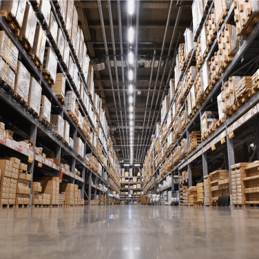 Wide-angle view of a large warehouse with tall shelves stacked with boxes, packages, and goods. The polished floor reflects the bright overhead lights, and the shelves extend into the distance.