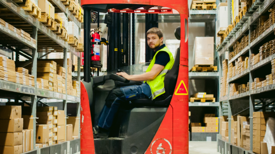 A man wearing a yellow safety vest drives a red forklift down an aisle in a warehouse, surrounded by shelves filled with cardboard boxes.