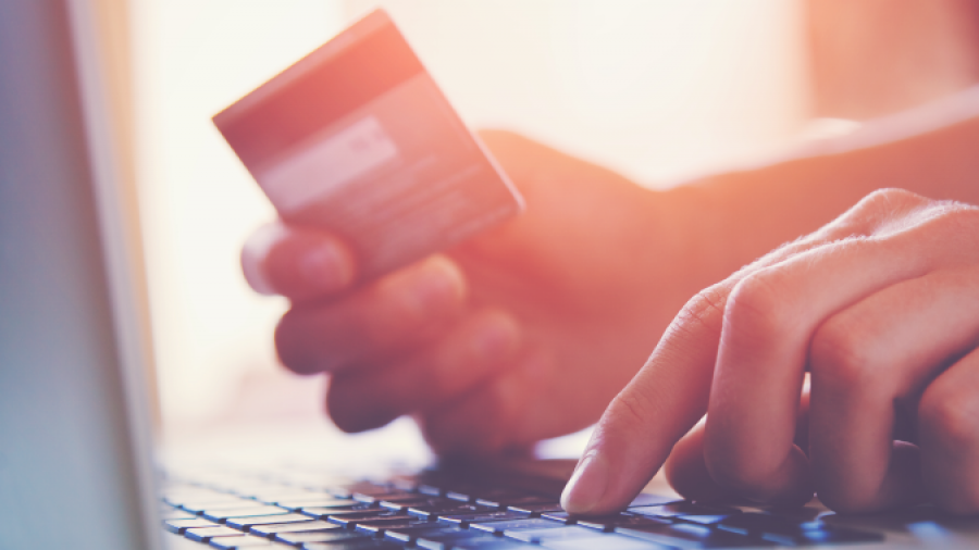 A person is holding a credit card and typing on a laptop keyboard, suggesting they are making an online purchase or payment. The image is softly lit with a warm tone.
