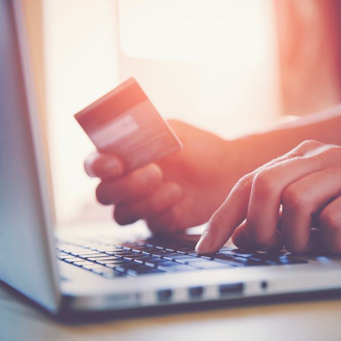 A person is holding a credit card and typing on a laptop keyboard, suggesting they are making an online purchase or payment. The image is softly lit with a warm tone.