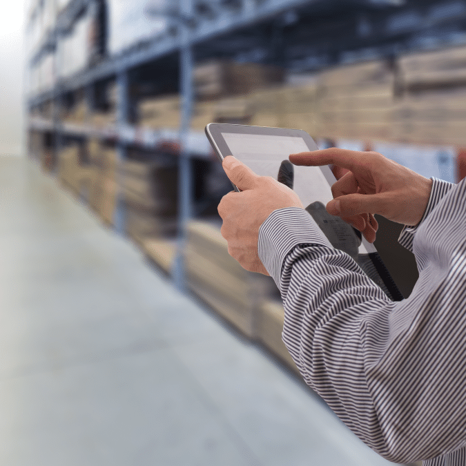 A person in business attire uses a tablet in a warehouse, showcasing distribution center automation, with shelves stocked with boxes in the background.