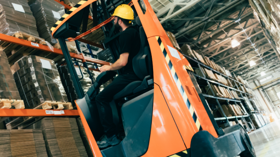 A worker in a yellow hard hat operates an orange forklift, lifting pallets inside a large warehouse with high shelves stacked with cardboard boxes.