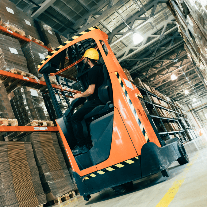 A worker in a yellow hard hat operates an orange forklift, lifting pallets inside a large warehouse with high shelves stacked with cardboard boxes.
