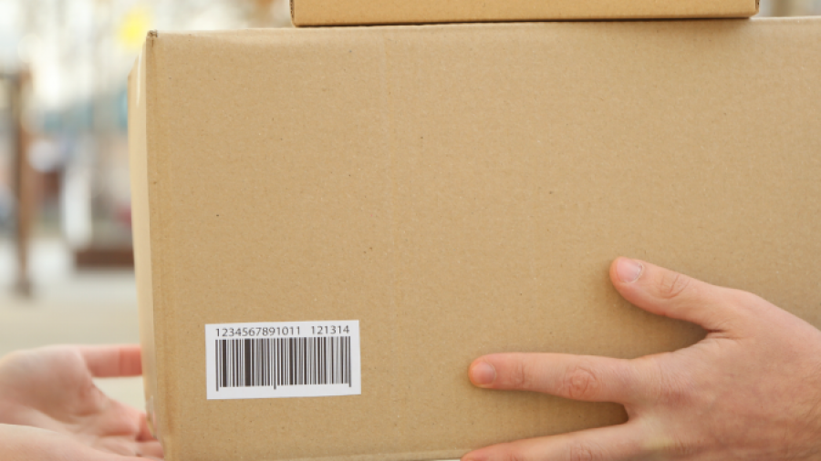 Two people exchange cardboard boxes outdoors; one person hands over two boxes with barcode labels to another person, who is receiving them with both hands. The background is blurred.