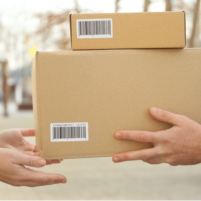 Two people exchange cardboard boxes outdoors; one person hands over two boxes with barcode labels to another person, who is receiving them with both hands. The background is blurred.