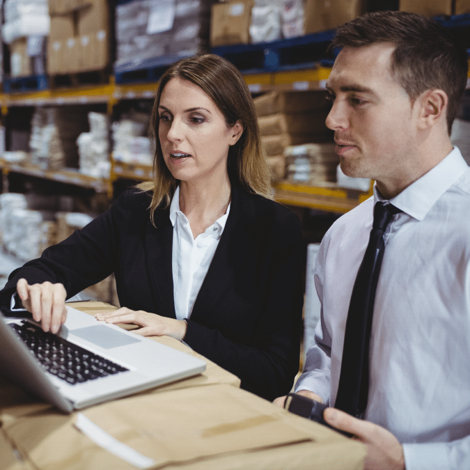 Two business professionals in formal attire look at a laptop on a desk in a warehouse, surrounded by shelves filled with boxes and packages.