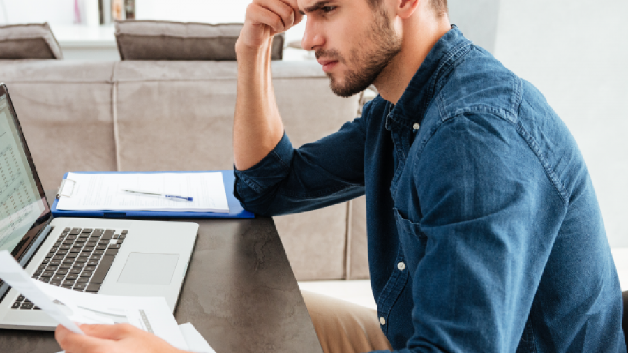 A man sits at a table, looking concerned while reviewing financial documents in front of a laptop, with charts and a pen on the table, in a home setting.