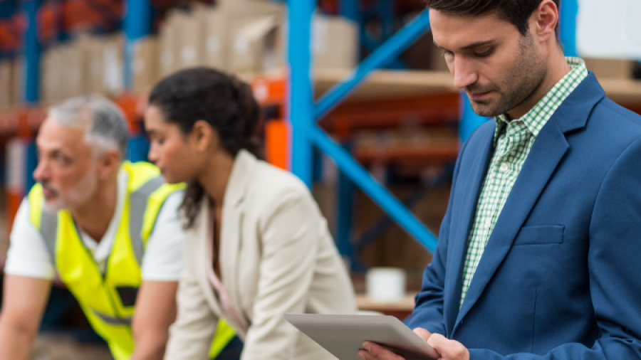 A man in a blue suit uses a tablet in a warehouse, supporting Lean Process Improvements, while two colleagues—one in a safety vest and one in business attire—review documents at a table. Shelves with boxes line the warehouse.