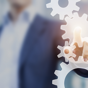 A person in a business suit is pressing a glowing gear icon on a transparent virtual touchscreen, symbolizing technology, Lean Process Improvements, or innovation. The blurred background emphasizes the focus on efficient process management.