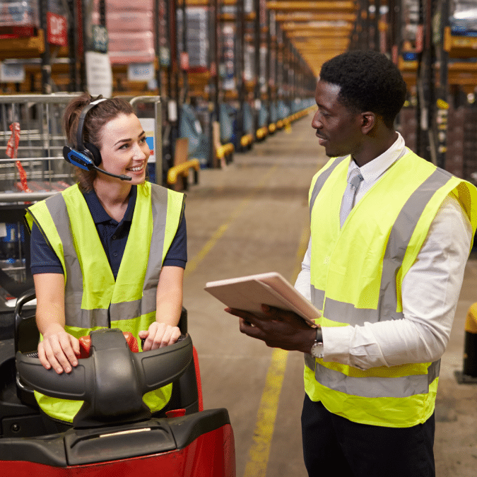 A woman in a safety vest and headset sits on a forklift, smiling and talking to a man in a safety vest holding a tablet in a warehouse aisle lined with shelves.