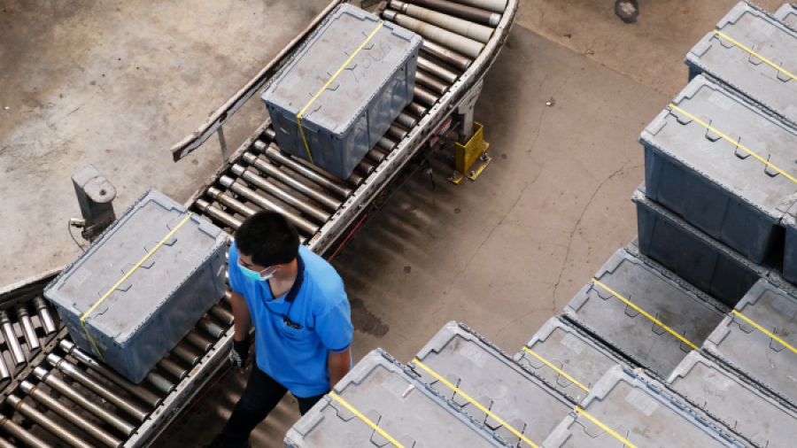 A person in a blue shirt and face mask walks beside a conveyor belt with large gray crates in an industrial warehouse setting. More crates are stacked neatly nearby.