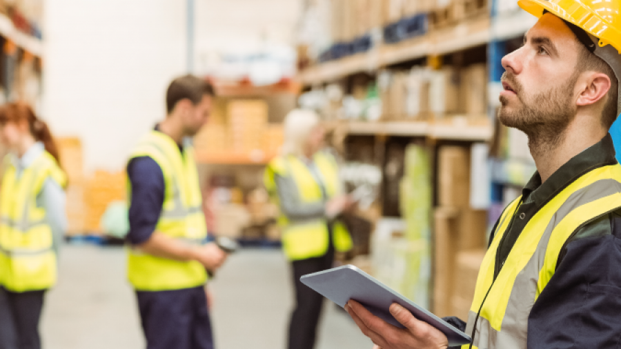 A man in a yellow safety vest and hard hat holds a tablet and looks up in a warehouse, while other workers in safety gear are seen working in the background among shelves of boxes.