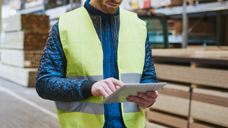 A worker in a yellow safety vest stands in a warehouse, using a tablet for warehouse optimization. He has a tool belt with tools, and stacks of wooden boards are visible in the background.