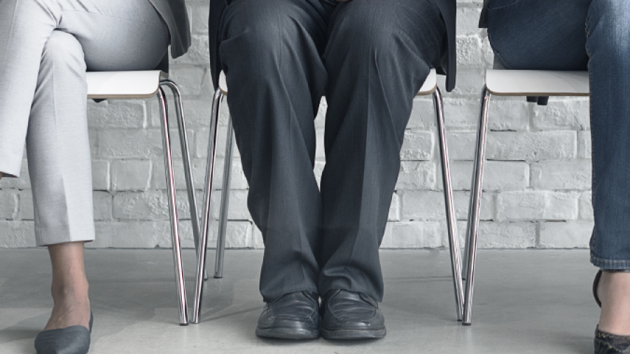 Three people in business attire sit on chairs against a white brick wall. They are holding phones or tablets, and only their bodies and legs are visible, not their faces.