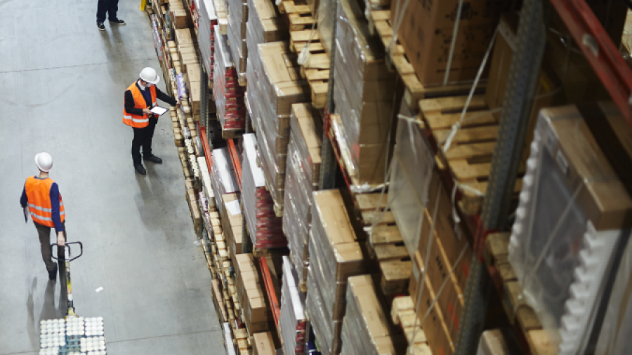 A warehouse with tall shelves stacked with boxes and three workers in safety vests and helmets. One worker is moving a pallet jack while two others are checking inventory on clipboards.
