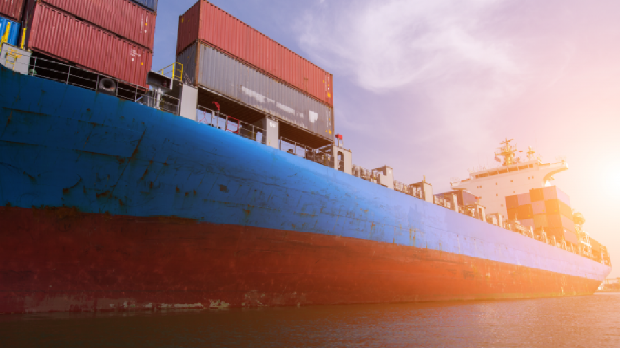 A large cargo ship loaded with colorful shipping containers sails on calm water at sunset, with the sun glowing in the background.
