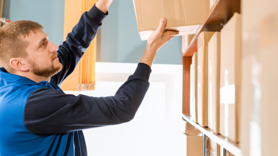A man in a blue vest reaches up to place or retrieve a cardboard box from a high shelf in a storage or warehouse setting. Several other boxes are visible on the shelves around him.
