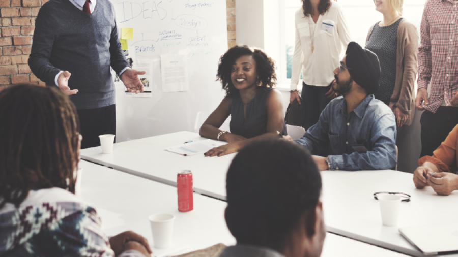 A diverse group of people are smiling and engaging in a meeting around a conference table, with one person standing and presenting ideas on a whiteboard in a modern office setting.