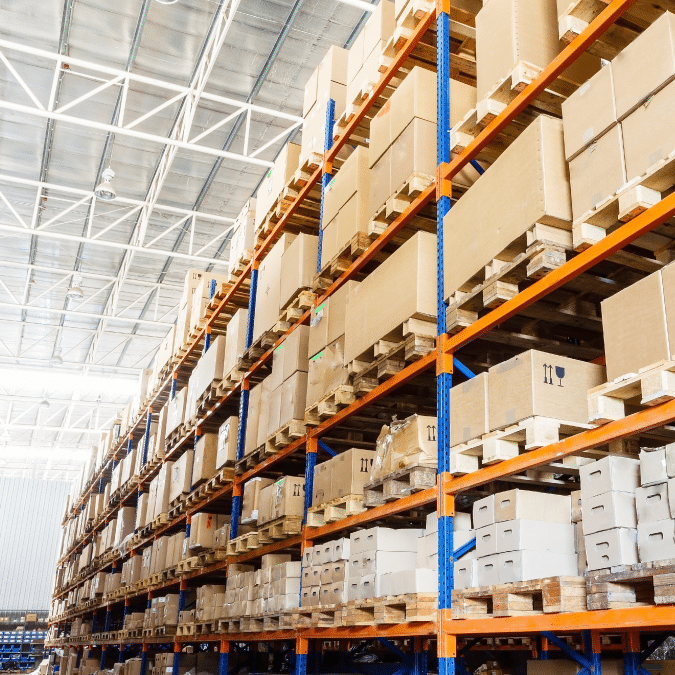 Large warehouse interior with tall shelves stacked with cardboard boxes and packages on wooden pallets. The space is brightly lit with natural and artificial light, and the shelves are organized and orderly.