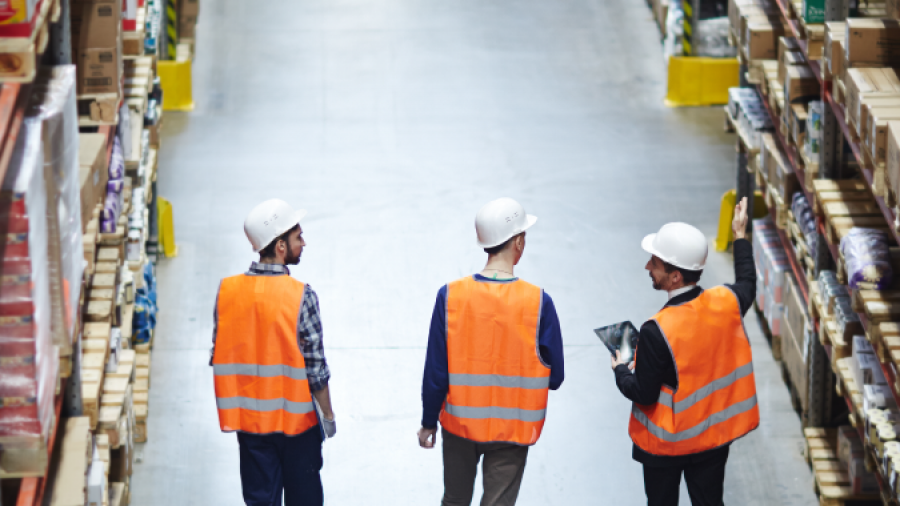 Three workers in safety vests and helmets walk down an aisle in a warehouse, surrounded by tall shelves stacked with boxes and goods. One worker gestures towards the shelves while another holds a tablet.