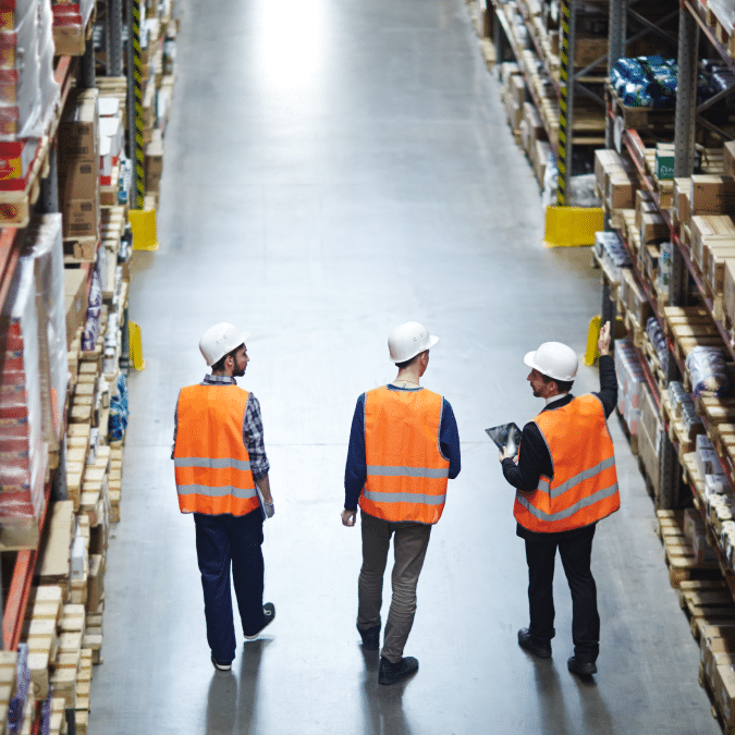 Three workers in safety vests and helmets walk down an aisle in a warehouse, surrounded by tall shelves stacked with boxes and goods. One worker gestures towards the shelves while another holds a tablet.