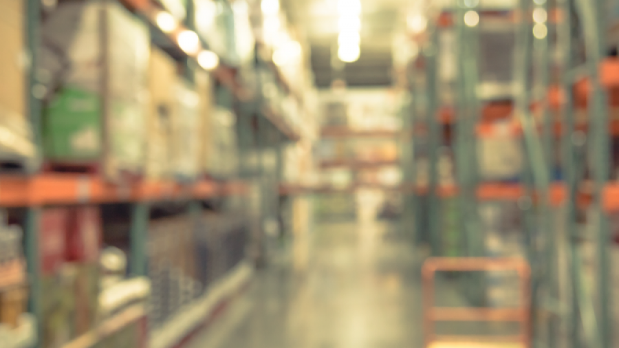 A blurred image of a warehouse aisle with shelves stacked with boxes and a trolley in the center, under bright overhead lighting.