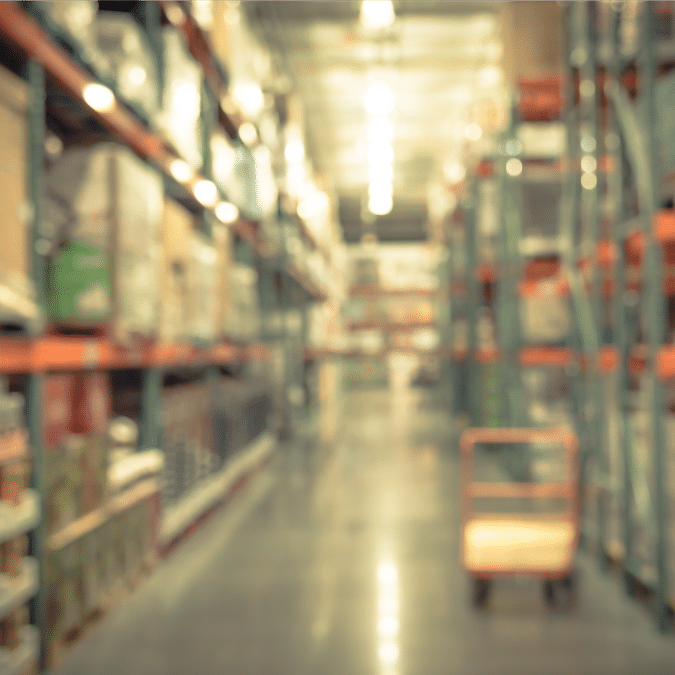 A blurred image of a warehouse aisle with shelves stacked with boxes and a trolley in the center, under bright overhead lighting.