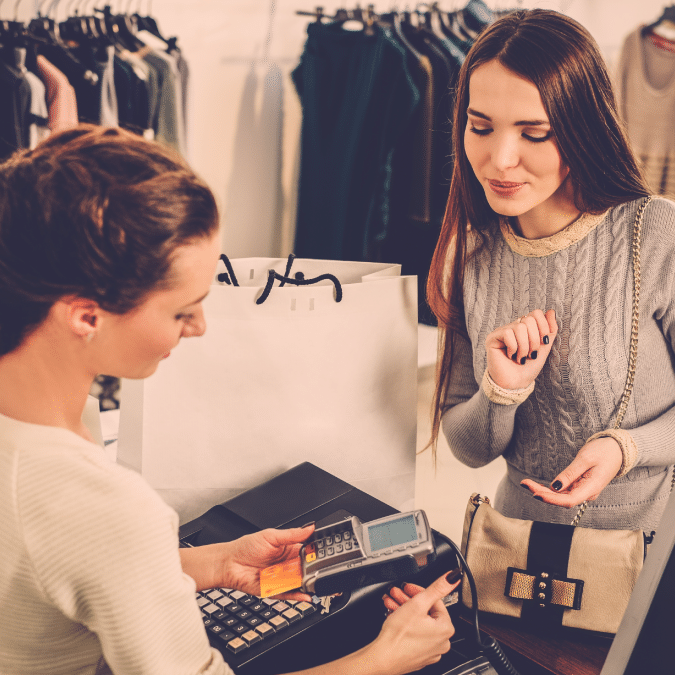 A woman at a clothing store counter smiles as a cashier prepares a card payment terminal. Shopping bags and clothing racks are visible in the background.