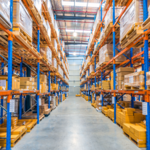 A large warehouse with tall shelves stacked with boxes and packages on both sides of a wide central aisle. The space is brightly lit with a high ceiling, organized storage, and efficient warehouse slotting optimization.