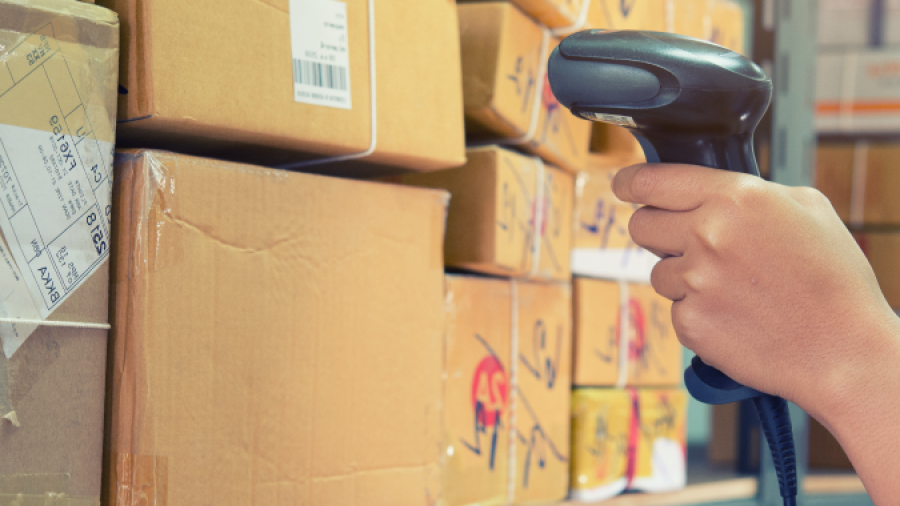A person scans a barcode on a cardboard box with a handheld scanner in a warehouse, surrounded by stacked packages on metal shelves.