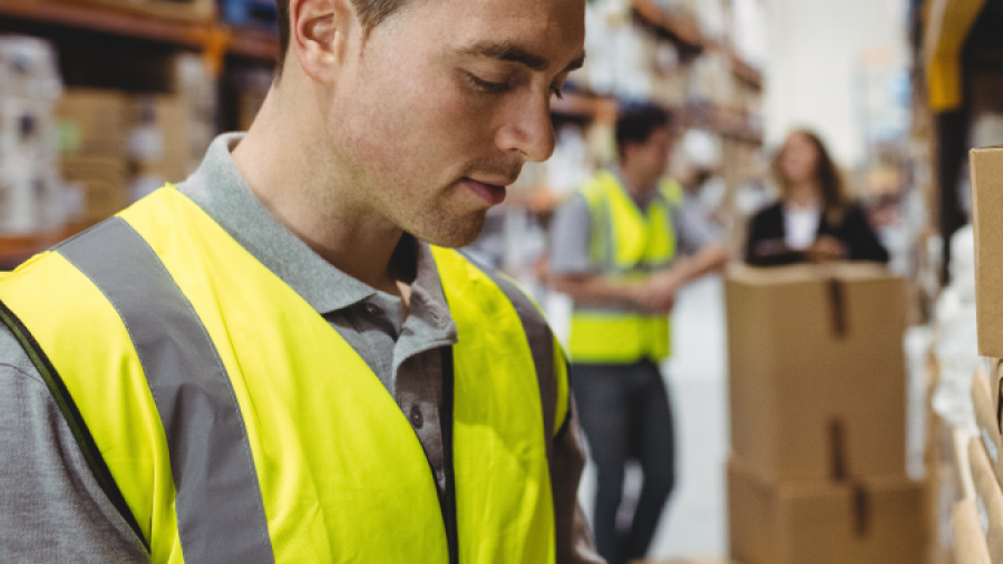 A warehouse worker wearing a yellow safety vest uses a handheld scanner to check inventory on shelves, with other workers and stacked boxes visible in the background.