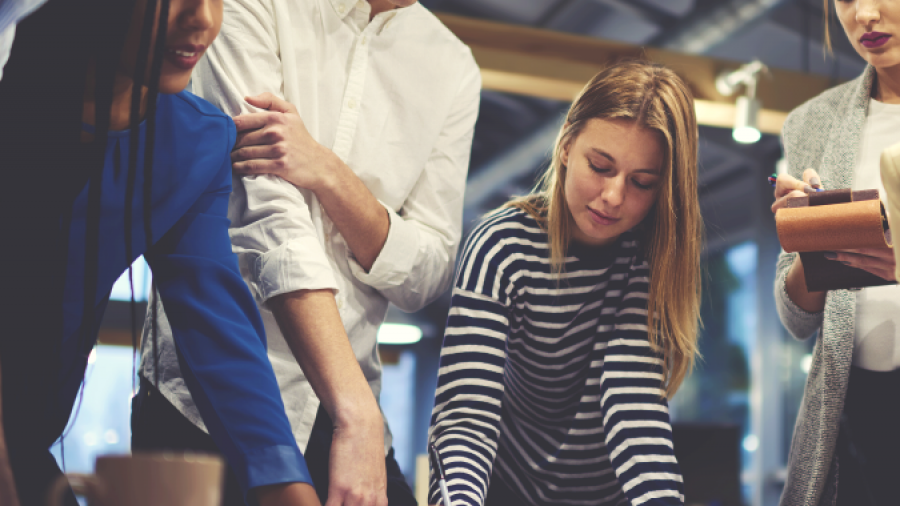 A group of young professionals collaborates around a table, looking at documents and taking notes in a modern office setting with warm lighting.