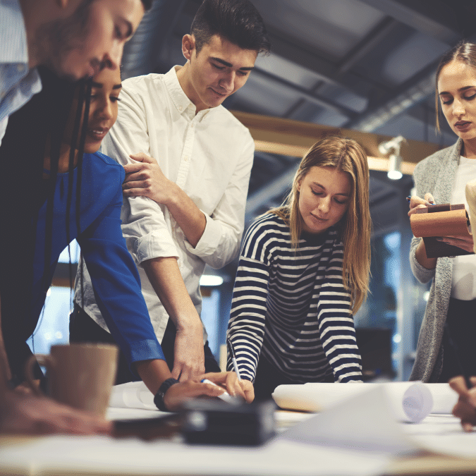 A group of young professionals collaborates around a table, looking at documents and taking notes in a modern office setting with warm lighting.