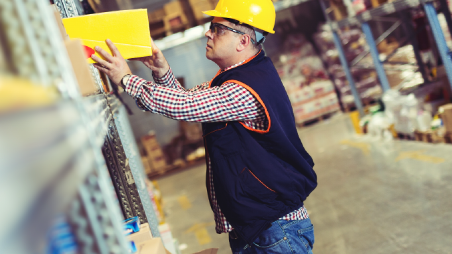 A worker wearing a yellow hard hat and safety vest reaches for a yellow box on a high shelf in a warehouse filled with boxes and supplies.