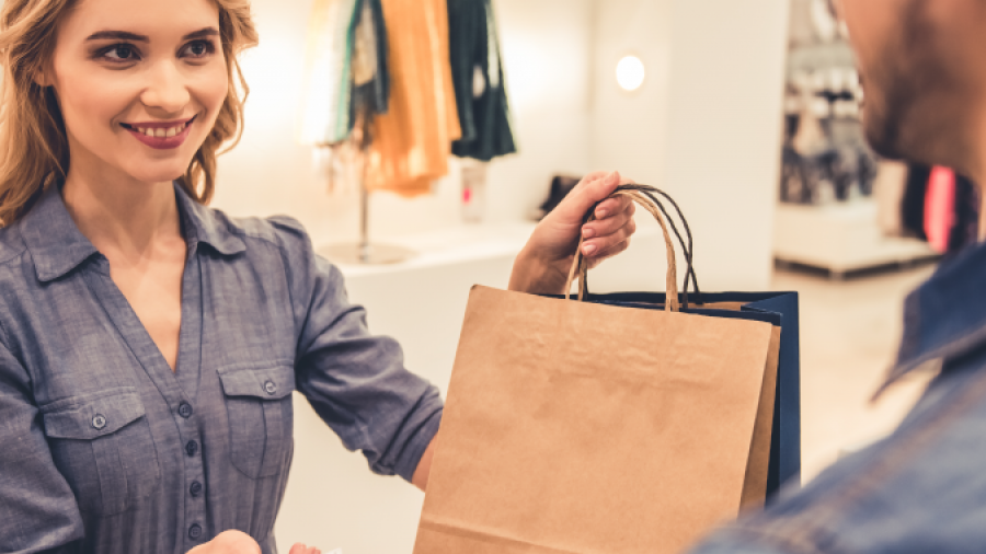 A smiling woman at a clothing store counter hands a paper shopping bag to a customer who is holding a credit card. Clothing racks and shelves are visible in the background.