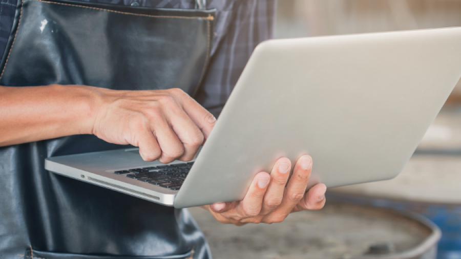 A person wearing a dark apron and plaid shirt uses a laptop, standing in what appears to be a workshop or industrial setting. Only the torso and hands are visible.