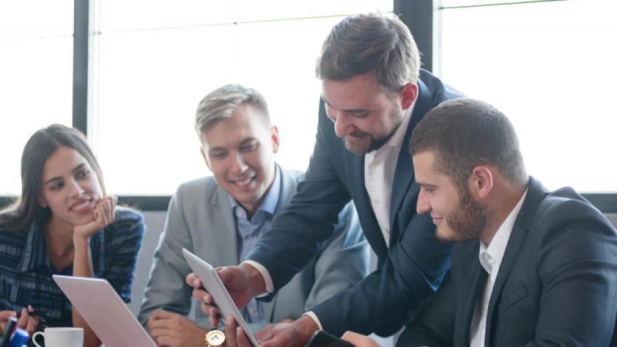 Four business professionals sit around a table in a bright office, collaborating and smiling as one man shows something on a tablet to the group. Laptops, papers, and fruit are on the table.