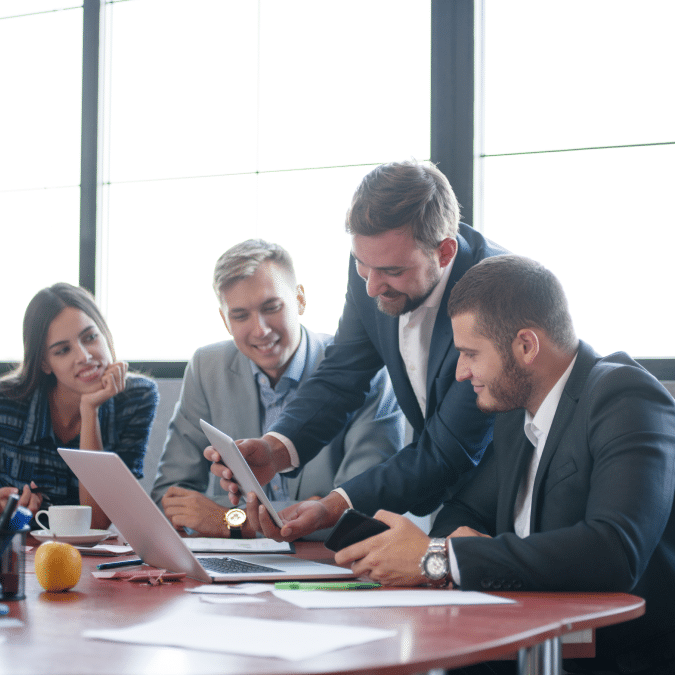 Four business professionals sit around a table in a bright office, collaborating and smiling as one man shows something on a tablet to the group. Laptops, papers, and fruit are on the table.