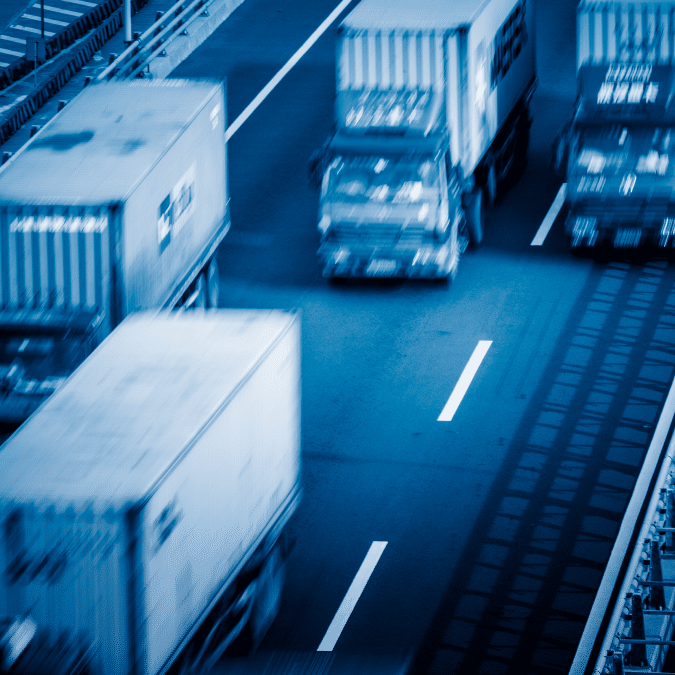 A blurred image of several large trucks driving quickly on a multi-lane highway, tinted blue to emphasize speed and motion—highlighting the efficiency offered by advanced transportation management systems.